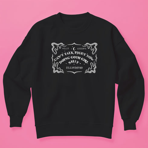 Black sweatshirt featuring white ouija board design with text "CAN'T TALK RIGHT NOW DOING GOTH GIRL SHIT". Ornate border surrounds the design, giving a mystical, witchy vibe.