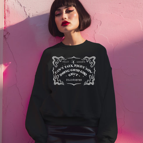 Black sweatshirt featuring white ouija board design with text "CAN'T TALK RIGHT NOW DOING GOTH GIRL SHIT". Ornate border surrounds the design, giving a mystical, witchy vibe.