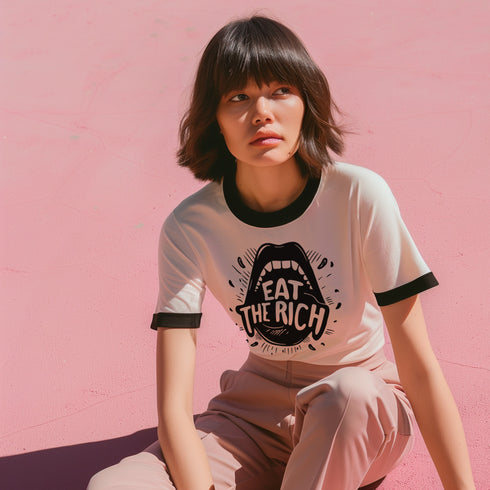 Heather gray ringer t-shirt with black contrast trim on the collar and sleeve cuffs. The front features a bold black graphic of an open mouth with the phrase "EAT THE RICH" in capital letters, integrated into the teeth. Surrounding the design are hand-drawn lines and accents, giving it a raw, punk-inspired aesthetic. The t-shirt has a vintage-inspired fit, made from soft cotton fabric with a slightly relaxed silhouette. The high-contrast black and gray design enhances the statement-making impact of the grap