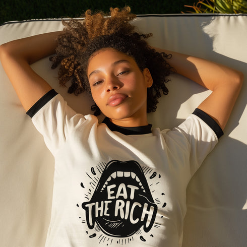 Heather gray ringer t-shirt with black contrast trim on the collar and sleeve cuffs. The front features a bold black graphic of an open mouth with the phrase "EAT THE RICH" in capital letters, integrated into the teeth. Surrounding the design are hand-drawn lines and accents, giving it a raw, punk-inspired aesthetic. The t-shirt has a vintage-inspired fit, made from soft cotton fabric with a slightly relaxed silhouette. The high-contrast black and gray design enhances the statement-making impact of the grap