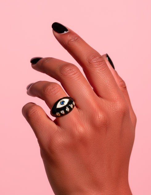 Close-up of a gold-plated Evil Eye Signet Ring in black enamel with micro-set zircon details, worn on a hand with black nails. The protective evil eye design sparkles under studio light against a soft pink background. Stylish occult jewelry by Honey Dazed for alt fashion lovers.