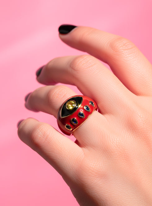 A red enamel Evil Eye Signet Ring with gold plating and zircon gemstone center, shown on a model’s hand with red nails. The ring’s symbolic design represents protection and empowerment. Bright yellow backdrop for modern maximalist appeal.