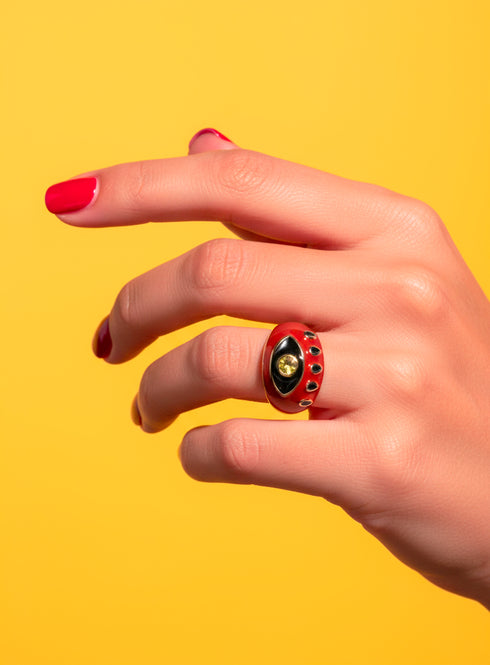 A red enamel Evil Eye Signet Ring with gold plating and zircon gemstone center, shown on a model’s hand with red nails. The ring’s symbolic design represents protection and empowerment. Bright yellow backdrop for modern maximalist appeal.