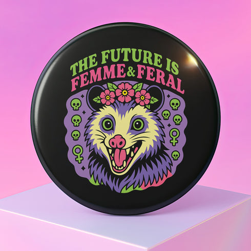 A round, black pinback button with a high-gloss finish features the text "THE FUTURE IS FEMME & FERAL" in a retro font of lime green and pastel pink. In the center, a screaming possum with wild eyes and a pink-lined mouth is illustrated in a pastel goth style with purple, yellow, and pink fur. The possum wears a pink flower crown. The illustration is framed by a circle of small skulls and pink Venus (female) symbols on a purple background. This is a piece of feminist possumcore flair.