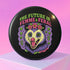 A round, black pinback button with a high-gloss finish features the text "THE FUTURE IS FEMME & FERAL" in a retro font of lime green and pastel pink. In the center, a screaming possum with wild eyes and a pink-lined mouth is illustrated in a pastel goth style with purple, yellow, and pink fur. The possum wears a pink flower crown. The illustration is framed by a circle of small skulls and pink Venus (female) symbols on a purple background. This is a piece of feminist possumcore flair.