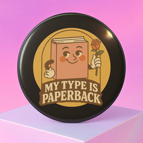 A glossy black round pin-back button with a retro 1970s-style design. The center shows a smiling cartoon paperback book holding a red rose, colored light brown with white gloves and feet. Below, “MY TYPE IS PAPERBACK” appears in chunky brown-and-tan 70s lettering. This bookish flair piece has a high-gloss, scratch-resistant mylar finish, secure safety pin backing, and comes in two- or three-inch sizes.
