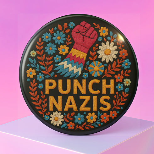 Round black pinback button with bold embroidered-style design featuring a raised red fist, zigzag sleeve, and colorful folk art flowers in orange, teal, yellow, and white. Center text reads “PUNCH NAZIS” in bright golden thread-style lettering. The design is bold, anti-fascist, and maximalist, shown on a glossy pin surface against a pink and purple gradient background. Statement accessory for protest fashion, punk style, and political activism.