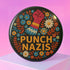 Round black pinback button with bold embroidered-style design featuring a raised red fist, zigzag sleeve, and colorful folk art flowers in orange, teal, yellow, and white. Center text reads “PUNCH NAZIS” in bright golden thread-style lettering. The design is bold, anti-fascist, and maximalist, shown on a glossy pin surface against a pink and purple gradient background. Statement accessory for protest fashion, punk style, and political activism.