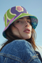 Reversible bucket hat shown on a white background with two distinct prints. One side is a flowing oceanic abstract with lilac and teal wavy stripes, mint green dots, and a royal blue base. The reverse is a retro mod geometric floral in dusty rose and muted purple petals outlined in cream and olive green. The hat has a structured flat crown, a downward sloping brim with visible stitching, and a textured woven finish. Maximalist art nouveau reversible bucket hat and psychedelic festival sun hat for quirky dop