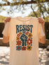 Cream unisex t-shirt featuring a centered design with dark teal raised fist surrounded by folk art botanical elements: crimson and red 5-petal daisies, golden yellow and orange flowers, cornflower blue petals, and small scattered wildflower accents arranged in a symmetrical medallion composition. Dark teal "RESIST" text appears above the fist. The design uses faux embroidery 2D print with dimensional texture detail. Shown on unisex fit with ribbed knit collar and short sleeves.