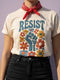 Cream unisex t-shirt featuring a centered design with dark teal raised fist surrounded by folk art botanical elements: crimson and red 5-petal daisies, golden yellow and orange flowers, cornflower blue petals, and small scattered wildflower accents arranged in a symmetrical medallion composition. Dark teal "RESIST" text appears above the fist. The design uses faux embroidery 2D print with dimensional texture detail. Shown on unisex fit with ribbed knit collar and short sleeves.