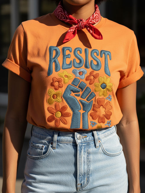 Cream unisex t-shirt featuring a centered design with dark teal raised fist surrounded by folk art botanical elements: crimson and red 5-petal daisies, golden yellow and orange flowers, cornflower blue petals, and small scattered wildflower accents arranged in a symmetrical medallion composition. Dark teal "RESIST" text appears above the fist. The design uses faux embroidery 2D print with dimensional texture detail. Shown on unisex fit with ribbed knit collar and short sleeves.