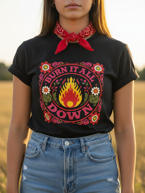 A high-resolution product image of a black unisex t-shirt featuring the ‘BURN IT ALL DOWN’ design in a folk-art style. The central motif is a vibrant orange and yellow flame surrounded by a circular medallion and a square floral border with red and pink flowers, white daisies, and green leaves. The print uses a faux-embroidery technique to create a trompe-l’œil 3D texture on a smooth 2D surface. The shirt has a ribbed knit collar, set against a high-contrast background.