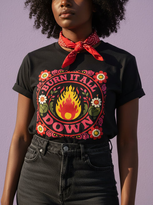A high-resolution product image of a black unisex t-shirt featuring the ‘BURN IT ALL DOWN’ design in a folk-art style. The central motif is a vibrant orange and yellow flame surrounded by a circular medallion and a square floral border with red and pink flowers, white daisies, and green leaves. The print uses a faux-embroidery technique to create a trompe-l’œil 3D texture on a smooth 2D surface. The shirt has a ribbed knit collar, set against a high-contrast background.