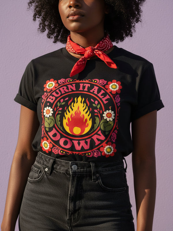 A high-resolution product image of a black unisex t-shirt featuring the ‘BURN IT ALL DOWN’ design in a folk-art style. The central motif is a vibrant orange and yellow flame surrounded by a circular medallion and a square floral border with red and pink flowers, white daisies, and green leaves. The print uses a faux-embroidery technique to create a trompe-l’œil 3D texture on a smooth 2D surface. The shirt has a ribbed knit collar, set against a high-contrast background.