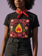 A high-resolution product image of a black unisex t-shirt featuring the ‘BURN IT ALL DOWN’ design in a folk-art style. The central motif is a vibrant orange and yellow flame surrounded by a circular medallion and a square floral border with red and pink flowers, white daisies, and green leaves. The print uses a faux-embroidery technique to create a trompe-l’œil 3D texture on a smooth 2D surface. The shirt has a ribbed knit collar, set against a high-contrast background.