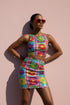 Form-fitting bodycon mini skirt with a mid-rise elastic waistband, falling to mid-thigh in smooth stretch fabric. It features a bold retro patchwork floral print: large mod daisies in red, purple, yellow, and green, set on a sky blue background with scattered green leaves. A central column frames single flowers on purple and pink tiles, edged by red and pink beaded borders. The all-over design is a flat dye-sublimated print, available in a smooth knit or glitter knit finish.