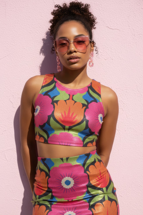 Form-fitting bodycon mini skirt with a mid-rise elastic waistband, falling to mid-thigh in smooth stretch fabric. It features a bold 70s pop art floral print: oversized hot pink flowers with cream scalloped centers, royal blue middles, and fine purple rays, beside a flaming orange tulip, royal blue geometric shapes, and olive green leaves on a black background. The all-over design is a flat dye-sublimated print in vibrant, high-contrast color, available in a smooth knit or glitter knit finish.