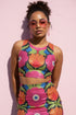 Form-fitting bodycon mini skirt with a mid-rise elastic waistband, falling to mid-thigh in smooth stretch fabric. It features a bold 70s pop art floral print: oversized hot pink flowers with cream scalloped centers, royal blue middles, and fine purple rays, beside a flaming orange tulip, royal blue geometric shapes, and olive green leaves on a black background. The all-over design is a flat dye-sublimated print in vibrant, high-contrast color, available in a smooth knit or glitter knit finish.