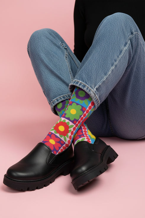 A pair of bright crew socks featuring a vibrant 70s-style maximalist patchwork design. The 2D flat print includes a collage of bold floral motifs in purple, lime green, red, and yellow set against alternating sky blue and light pink square panels. Features a red and white sawtooth border at the cuff and a vertical red scalloped stripe. Reinforced solid black toe and heel. High-quality accessory from black owned shops.