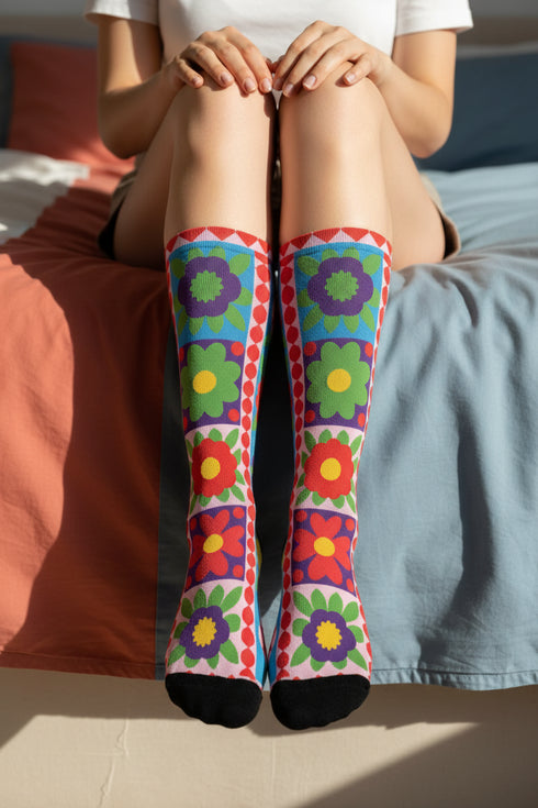 A pair of bright crew socks featuring a vibrant 70s-style maximalist patchwork design. The 2D flat print includes a collage of bold floral motifs in purple, lime green, red, and yellow set against alternating sky blue and light pink square panels. Features a red and white sawtooth border at the cuff and a vertical red scalloped stripe. Reinforced solid black toe and heel. High-quality accessory from black owned shops.