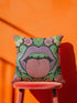 A square decorative pillow cover featuring a flat print design that mimics textured embroidery. The bold motif is set on a solid black background and centers on a pair of large, light purple lips with a bright pink, textured-looking tongue sticking out. Green ruffled leaves outline the mouth, and stylized pink daisy-like flowers sit at the top corners and bottom edges. The fabric has a faux linen weave texture, presenting a vibrant, symmetrical, and maximalist pop art aesthetic.