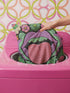 A square decorative pillow cover featuring a flat print design that mimics textured embroidery. The bold motif is set on a solid black background and centers on a pair of large, light purple lips with a bright pink, textured-looking tongue sticking out. Green ruffled leaves outline the mouth, and stylized pink daisy-like flowers sit at the top corners and bottom edges. The fabric has a faux linen weave texture, presenting a vibrant, symmetrical, and maximalist pop art aesthetic.