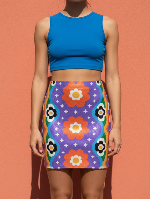 Form-fitting bodycon mini skirt with a mid-rise elastic waistband, falling to mid-thigh in smooth stretch fabric. It features a bold groovy mod floral print: oversized orange flowers with white inner petals and golden yellow centers, black flower accents, and white four-point sparkles, all set on a deep purple background. Rainbow borders in blue, green, orange, and yellow run down each side. The all-over design is a flat dye-sublimated print, available in a smooth knit or glitter knit finish.