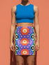 Form-fitting bodycon mini skirt with a mid-rise elastic waistband, falling to mid-thigh in smooth stretch fabric. It features a bold groovy mod floral print: oversized orange flowers with white inner petals and golden yellow centers, black flower accents, and white four-point sparkles, all set on a deep purple background. Rainbow borders in blue, green, orange, and yellow run down each side. The all-over design is a flat dye-sublimated print, available in a smooth knit or glitter knit finish.