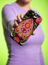 Black phone case featuring a faux embroidery style print with a central pink planchette design. The planchette contains a single eye motif and a circular sun element above it. Surrounding the design are orange, red, and pink flowers, green leaves, small cream stars, and crescent moons. The print creates a stitched textile illusion on a smooth matte protective case with dual layer construction.