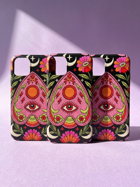 Black phone case featuring a faux embroidery style print with a central pink planchette design. The planchette contains a single eye motif and a circular sun element above it. Surrounding the design are orange, red, and pink flowers, green leaves, small cream stars, and crescent moons. The print creates a stitched textile illusion on a smooth matte protective case with dual layer construction.