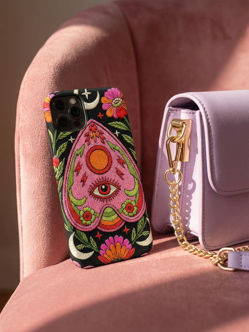 Black phone case featuring a faux embroidery style print with a central pink planchette design. The planchette contains a single eye motif and a circular sun element above it. Surrounding the design are orange, red, and pink flowers, green leaves, small cream stars, and crescent moons. The print creates a stitched textile illusion on a smooth matte protective case with dual layer construction.
