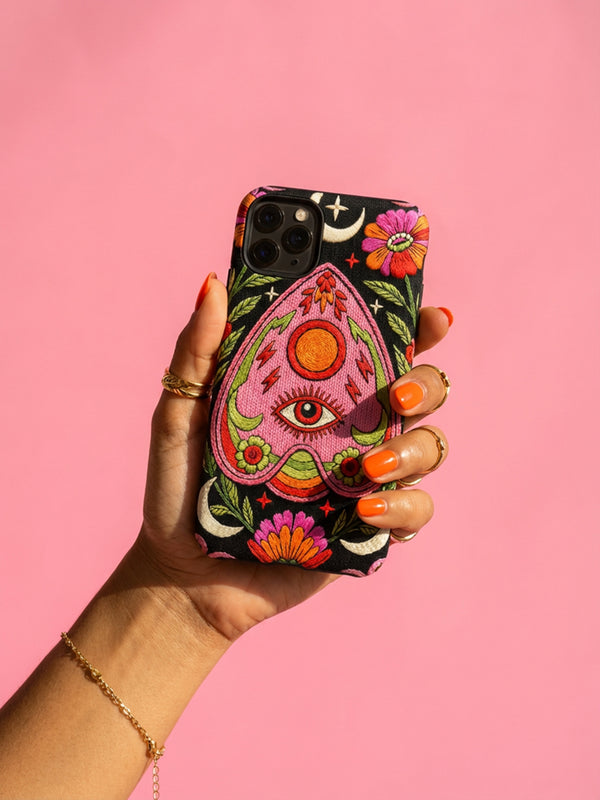 Black phone case featuring a faux embroidery style print with a central pink planchette design. The planchette contains a single eye motif and a circular sun element above it. Surrounding the design are orange, red, and pink flowers, green leaves, small cream stars, and crescent moons. The print creates a stitched textile illusion on a smooth matte protective case with dual layer construction.