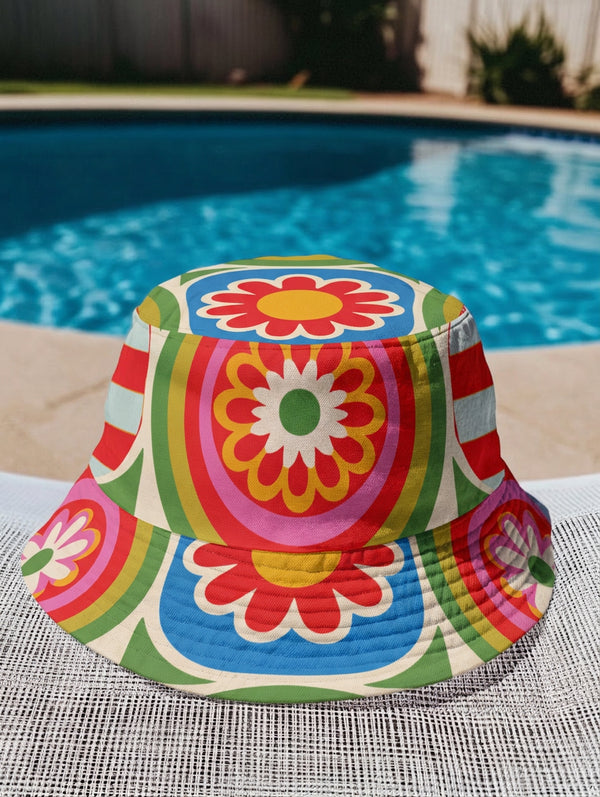 Reversible all-over print unisex bucket hat featuring two distinct bold designs. Side one showcases a psychedelic wavy stripe pattern in deep red, orange, green, blue, cream, and blush. Side two features a retro 70s groovy floral medallion print with large daisy motifs, red petals, yellow centers, and concentric pink, orange, and green rings on a cream background. Made from 100% polyester with a soft linen-feel texture. Moisture-wicking and breathable. Unisex sizing: XS/S and M/L/XL.
