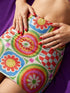 A vibrant bodycon mini skirt featuring a structured grid of retro geometric medallions. The pattern includes concentric circles in red, yellow, and blue, stylized daisy flower motifs, and red and white checkered squares. The skirt has a high-waist elastic band and a smooth, matte performance knit texture. The background is a neutral off-white. This maximalist design utilizes saturated primary colors to create a 70s-inspired psychedelic aesthetic. No purple or navy is present in the print.