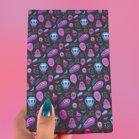 Spooky birthday wrapping paper with neon pink skulls, blue coffins, eyeballs, and bats on black background. Vibrant and gothic design for unique gift presentation.