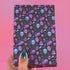 Spooky birthday wrapping paper with neon pink skulls, blue coffins, eyeballs, and bats on black background. Vibrant and gothic design for unique gift presentation.