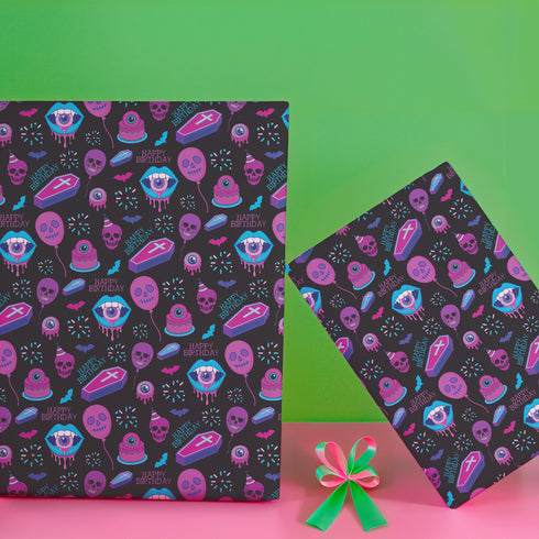 Spooky birthday wrapping paper with neon pink skulls, blue coffins, eyeballs, and bats on black background. Vibrant and gothic design for unique gift presentation.