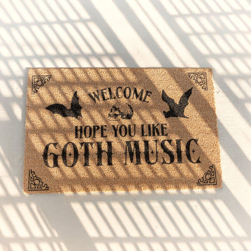 Tan coir doormat featuring "Welcome, Hope You Like Goth Music" in black text, with bat and skull imagery and gothic-style corner decorations.