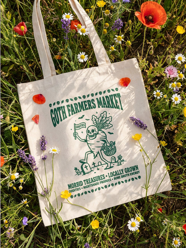 A natural beige canvas-textured tote bag featuring a forest green graphic illustration. The design depicts a skeleton gardener walking amongst tombstones, carrying a basket of vegetables and holding a flag that reads "Oddities." The main text reads "Goth Farmers Market" in vintage typography, with subtitles "Morbid Treasures" and "Locally Grown." The border features skulls and leaves. The bag has long handles and a flat profile, ideal for goth gardening and spooky plant lover gifts.