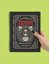 An 8.5 x 11 inch black hardcover spiral planner with a durable satin matte laminated finish. The front cover features a vintage-style white ornamental border, creating a gothic, macabre aesthetic. Inside the frame, the title "Calendar of Doom" is printed in an antique serif font. This undated weekly and monthly agenda is shown with a metal wire-o spiral binding on the right side. The design uses a black, white, and red color scheme, making it a unique piece of dark humor stationery