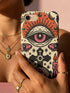 A vertical phone case design featuring a faux crochet texture illusion printed on a cream background. The symmetrical pattern centers on a large black eye with a pink iris and rust-orange sunburst lashes at the top. Below the eye is a black cross surrounded by small black stars. The bottom features a skull with a third rust-orange eye on its forehead. Flanking the skull are two candles with orange flames and rust-colored leaves. The design mimics the look of stitched yarn but is a 2d print.