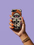 A vertical phone case design featuring a faux crochet texture illusion printed on a cream background. The symmetrical pattern centers on a large black eye with a pink iris and rust-orange sunburst lashes at the top. Below the eye is a black cross surrounded by small black stars. The bottom features a skull with a third rust-orange eye on its forehead. Flanking the skull are two candles with orange flames and rust-colored leaves. The design mimics the look of stitched yarn but is a 2d print.