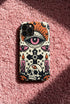 A vertical phone case design featuring a faux crochet texture illusion printed on a cream background. The symmetrical pattern centers on a large black eye with a pink iris and rust-orange sunburst lashes at the top. Below the eye is a black cross surrounded by small black stars. The bottom features a skull with a third rust-orange eye on its forehead. Flanking the skull are two candles with orange flames and rust-colored leaves. The design mimics the look of stitched yarn but is a 2d print.