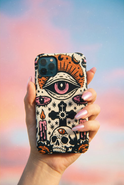 A vertical phone case design featuring a faux crochet texture illusion printed on a cream background. The symmetrical pattern centers on a large black eye with a pink iris and rust-orange sunburst lashes at the top. Below the eye is a black cross surrounded by small black stars. The bottom features a skull with a third rust-orange eye on its forehead. Flanking the skull are two candles with orange flames and rust-colored leaves. The design mimics the look of stitched yarn but is a 2d print.