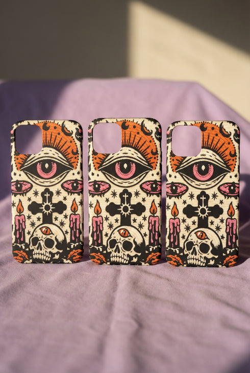 A vertical phone case design featuring a faux crochet texture illusion printed on a cream background. The symmetrical pattern centers on a large black eye with a pink iris and rust-orange sunburst lashes at the top. Below the eye is a black cross surrounded by small black stars. The bottom features a skull with a third rust-orange eye on its forehead. Flanking the skull are two candles with orange flames and rust-colored leaves. The design mimics the look of stitched yarn but is a 2d print.