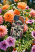 A vertical phone case design featuring a faux crochet texture illusion printed on a cream background. The symmetrical pattern centers on a large black eye with a pink iris and rust-orange sunburst lashes at the top. Below the eye is a black cross surrounded by small black stars. The bottom features a skull with a third rust-orange eye on its forehead. Flanking the skull are two candles with orange flames and rust-colored leaves. The design mimics the look of stitched yarn but is a 2d print.