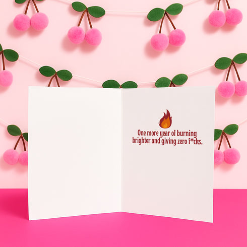 5x7 inch birthday greeting card featuring a bold psychedelic design with a snake wrapped around a melting candle, flames, mushrooms, and planets in red, orange, and pink tones. The card includes a white envelope and an interior message that reads, "One more year of burning brighter and giving zero f*cks." Printed on glossy silk stock.