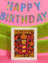 5x7 inch birthday greeting card featuring a bold psychedelic design with a snake wrapped around a melting candle, flames, mushrooms, and planets in red, orange, and pink tones. The card includes a white envelope and an interior message that reads, "One more year of burning brighter and giving zero f*cks." Printed on glossy silk stock.