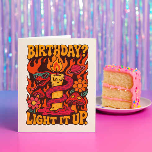 5x7 inch birthday greeting card featuring a bold psychedelic design with a snake wrapped around a melting candle, flames, mushrooms, and planets in red, orange, and pink tones. The card includes a white envelope and an interior message that reads, "One more year of burning brighter and giving zero f*cks." Printed on glossy silk stock.